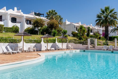 Piscina ensolarada em AlgarStays Alporchichos com água turquesa, espreguiçadeiras, apartamentos brancos, palmeiras e vistas para o mar