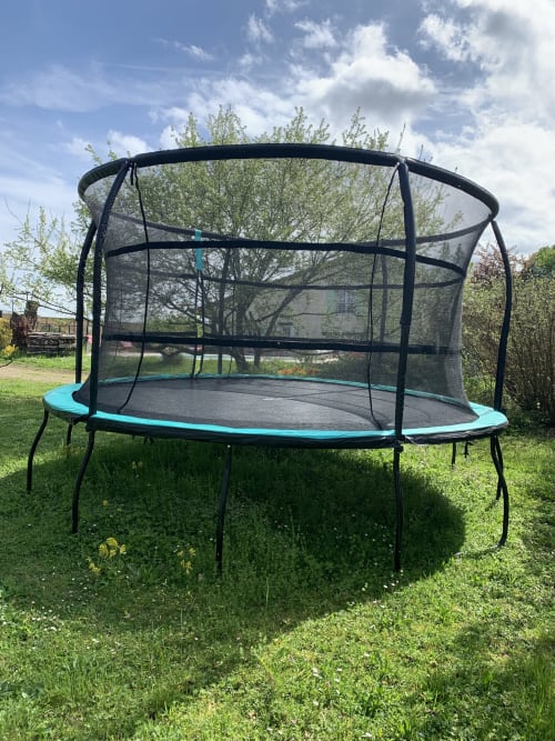 Trampoline bleu et noir avec filet de sécurité dans le jardin herbeux de la villa Maison Héléna, Lain, France