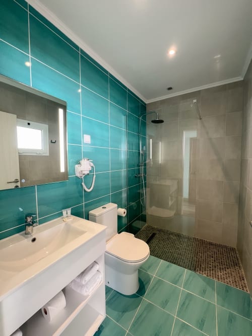 Banheiro moderno com azulejos turquesa, pia branca, vaso sanitário e chuveiro aberto no hotel Monte Horeb, Nadadouro