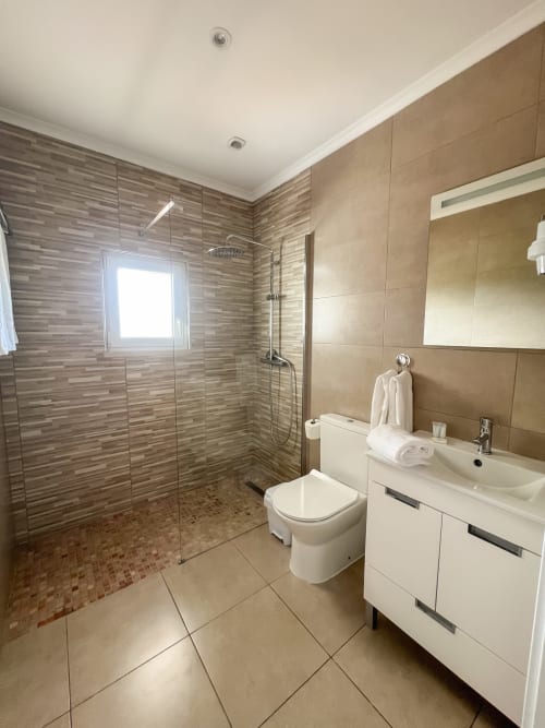 Banheiro moderno do Alojamento Local Monte Horeb com chuveiro de grande formato, bancada branca, sanitário e piso em cerâmica bege