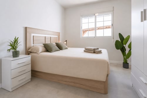 Dormitorio luminoso y accesible en Studios Sanchis, Alicante con cama de madera, mesita blanca, plantas en maceta y gran ventana