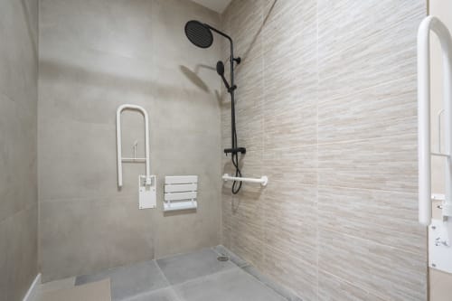 Baño accesible en Studios Sanchis Alicante con alcachofa de lluvia negra, barras de seguridad blancas y paredes de baldosas beige