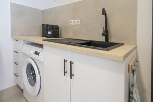Moderno lavadero en estudio hotelero de Alicante con lavadora, lavabo negro, encimera de madera y muebles blancos