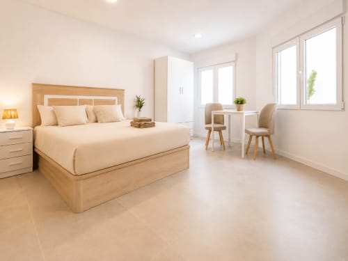 Dormitorio moderno en Studios Sanchis Alicante con cama de madera, escritorio, sillas y grandes ventanales con vistas a la piscina