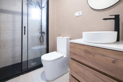 Baño moderno en Studios Sanchis Alicante con lavabo blanco de cerámica, grifo negro, inodoro blanco y ducha con mampara de cristal