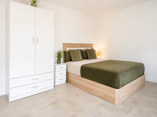 Moderno dormitorio estudio en Studios Sanchis Alicante con cama de madera, ropa de cama verde, armario blanco, plantas en maceta y