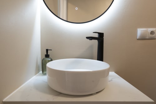 Baño moderno en Studios Sanchis Alicante: lavabo redondo blanco con grifo negro alto, espejo redondo iluminado, y