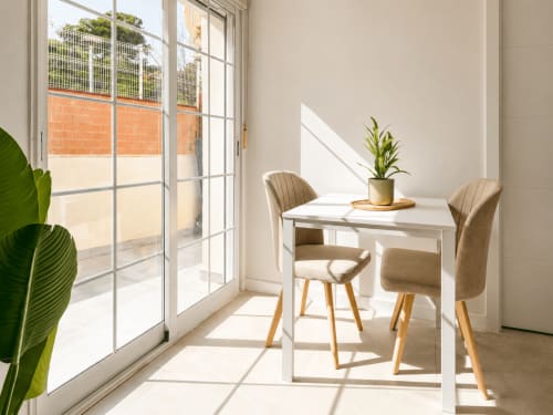Luminosa terraza de estudio en Studios Sanchis con mesa blanca, sillas beige, plantas en maceta y puertas de cristal con vistas a Alicante