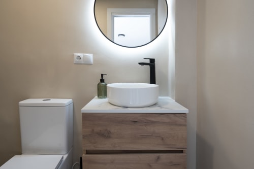 Baño moderno en Studios Sanchis Alicante con lavamanos redondo blanco, grifo negro, espejo iluminado, inodoro y elementos de madera