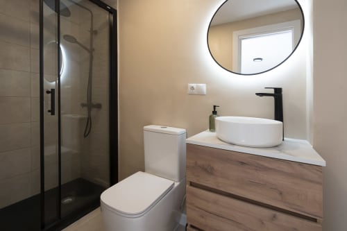 Baño moderno en el hotel Studios Sanchis Alicante con lavamanos tipo vasija, espejo redondo, grifo negro, inodoro y ducha abierta