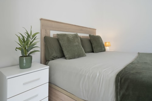 Dormitorio moderno en Studios Sanchis Alicante con cama matrimonial, almohadas verdes, mesilla blanca con planta e iluminación cálida