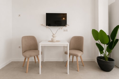 Zona de estar moderna en Studios Sanchis en Alicante con dos sillas beige, mesa blanca, TV montada en pared y planta decorativa