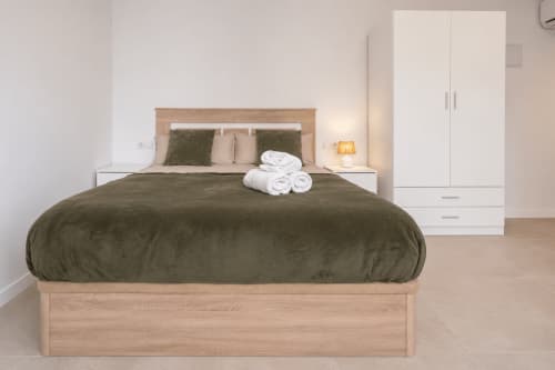 Moderno dormitorio en estudios de Studios Sanchis, Alicante con edredón oliva, marco de cama de madera, mesillas blancas, toallas enrolladas