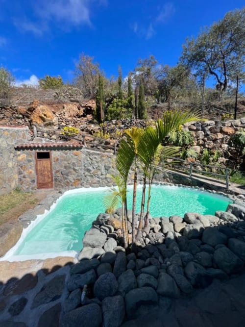 Piscina turquesa rodeada de rocas negras en Villa Awara, La Palma, con edificio de piedra y palmeras bajo cielo azul