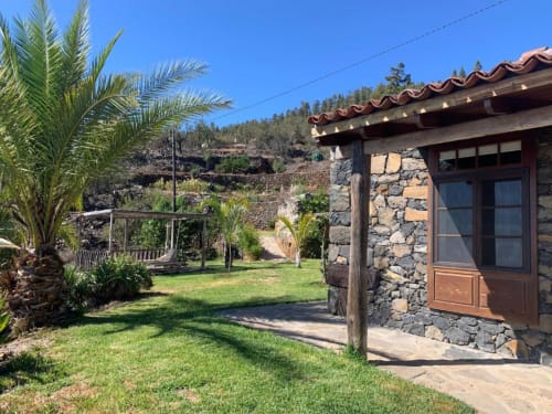 Villa de piedra con terraza en Rural La Palma, El Pinillo, con palmera, patio ajardinado y vistas montañosas bajo cielo azul despejado
