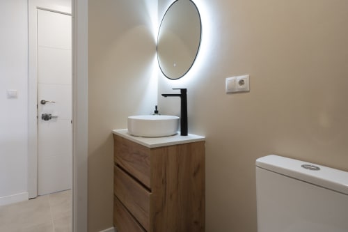 Baño moderno en Studios Sanchis Alicante con espejo iluminado redondo, lavamanos blanco, tocador de madera y grifo negro