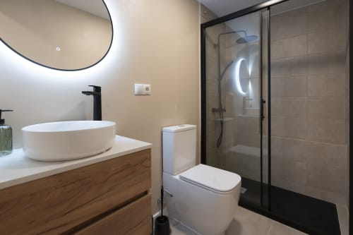 Baño moderno en Studios Sanchis Alicante con lavabo blanco, ducha abierta y mueble de madera