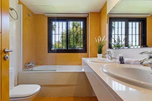 Modern bathroom with yellow tile walls, dual sinks, bathtub with shower, windows overlooking gardens at Ayma Villa in