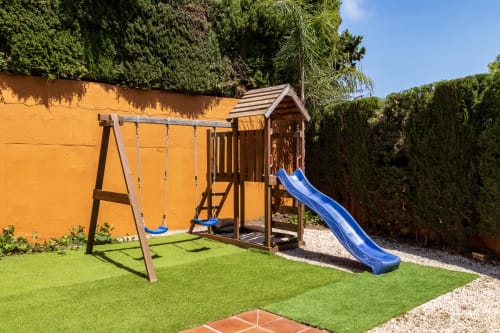 Backyard play area at Ayma Villa in Benalmádena with swing set, wooden playhouse, and blue slide on green lawn