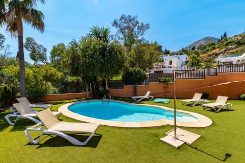 Luxury villa in Benalmádena with oval blue pool, white loungers on green lawn, and mountain views surrounded by orange walls