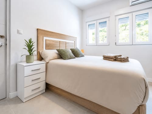 Moderno dormitorio en Studios Sanchis de Alicante con cama queen, mesita blanca, cabecero de madera, planta en maceta y luminoso