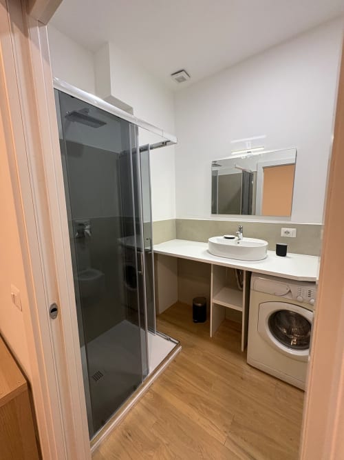Bagno moderno nell'appartamento ESCAPE Milano con doccia, lavandino, lavatrice, specchio e pavimento in legno