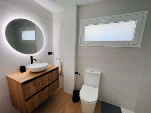 Baño moderno en apartamento La Restinga con espejo iluminado redondo, lavamanos blanco, tocador de madera e inodoro blanco