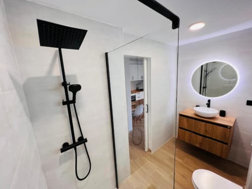 Baño moderno con ducha tipo lluvia negra, espejo redondo iluminado, tocador de madera y cabina de ducha de vidrio