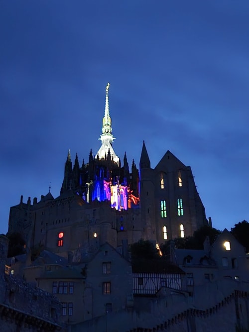 Mont-Saint-Michel illuminé au crépuscule avec des éclairages bleus et roses, abbaye gothique iconique et flèche contre un ciel crépusculaire