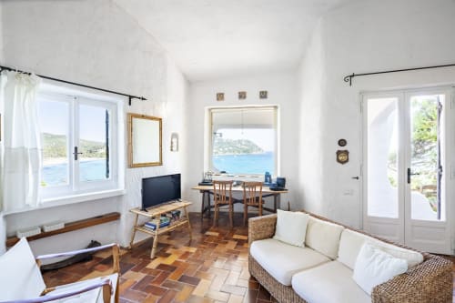 Luminoso soggiorno di villa sarda con vista mare, divano in vimini, scrivania in legno e paesaggio costiero attraverso ampie finestre