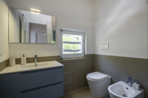 Bagno moderno presso Top Villas Sardegna con rivestimenti in piastrelle scure, lavabo, toilette, bidet e finestra a Quartu Sant'Elena
