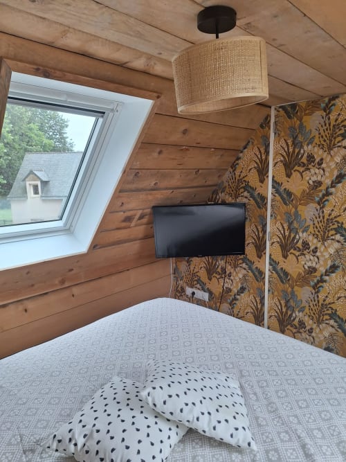 Cozy cottage bedroom at Le Village Du Moulin with wooden beams, wall-mounted TV, patterned wallpaper, and skylight window