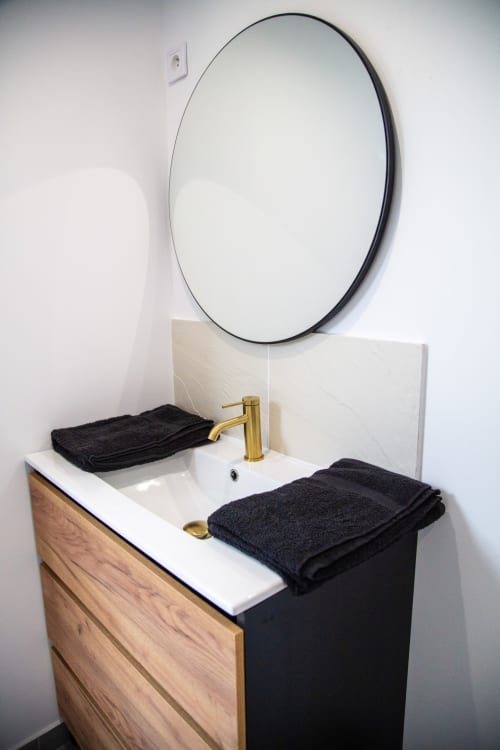 Vasque blanche moderne avec robinet doré, serviettes noires, meuble en bois et miroir rond à BALI ROOM