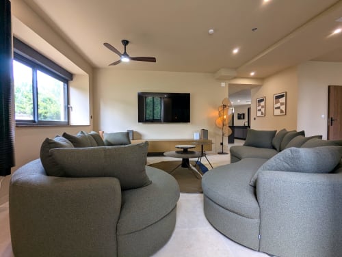 Salon moderne du Domaine Alga avec canapés gris arrondis, téléviseur écran plat, ventilateur de plafond et lumière naturelle depuis les grandes fenêtres