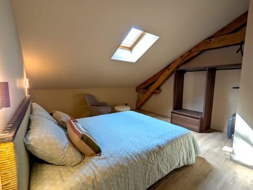 Chambre lumineuse sous les toits du Domaine Alga avec couvre-lit bleu, poutres apparentes, verrière et mobilier bois
