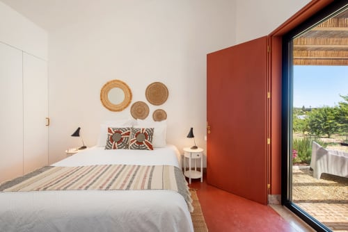 Quarto moderno em Monte das Açoteias com cama branca, parede de destaque em terracota, decoração com cestos tecidos e vista para o jardim em Tavira