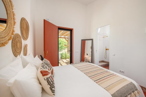 Quarto brilhante em Monte das Açoteias com roupas de cama brancas, cestos tran çados decorativos, acentos vermelhos e vistas para o jardim