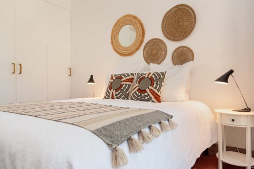 Quarto ensolarado em Monte das Açoteias com cama branca, cestos tramados na parede, almofadas estampadas e luminária preta moderna