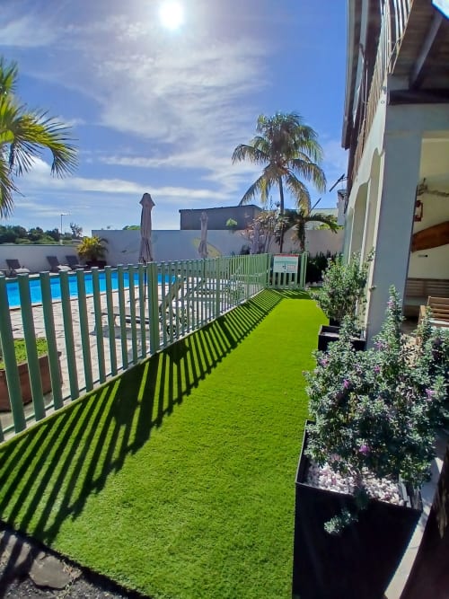 Sunny garden terrace at Ti Village Créole with green lawn, flowering plants, pool view, and palm trees in Sainte-Anne