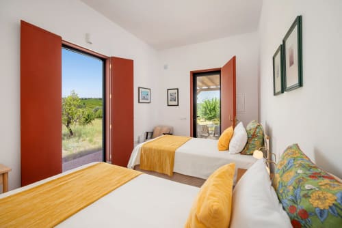 Quarto iluminado em Monte das Açoteias com duas camas, roupas de cama douradas, portas vermelhas com vista para o jardim e rústico Tavira