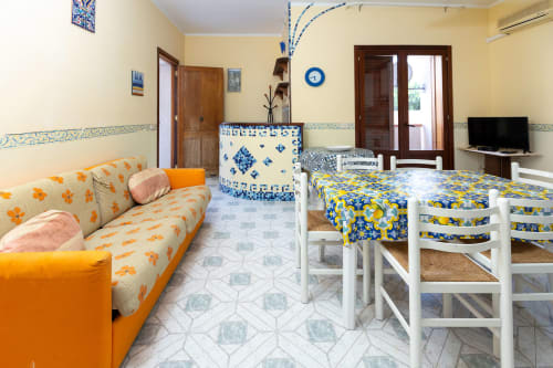 Luminoso appartamento a San Vito Lo Capo con divano arancione, tocchi blu, zona pranzo, TV e arredamento siciliano tradizionale