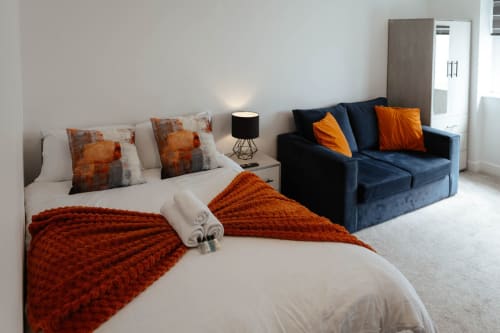 Modern studio apartment bedroom with orange accents, bed with decorative pillows, dark blue sofa, and city views at The Snug,