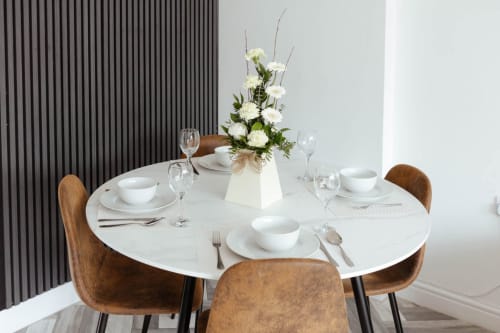 Elegant dining setup at JS Apartments Blackburn with round white table, brown chairs, white place settings, and white flower