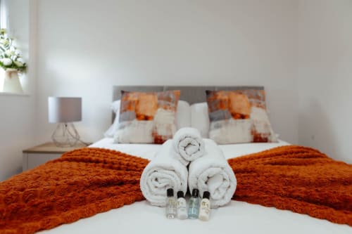 Welcoming bedroom at JS Apartments, Blackburn, with rolled white towels, toiletries, orange throws, and patterned pillows