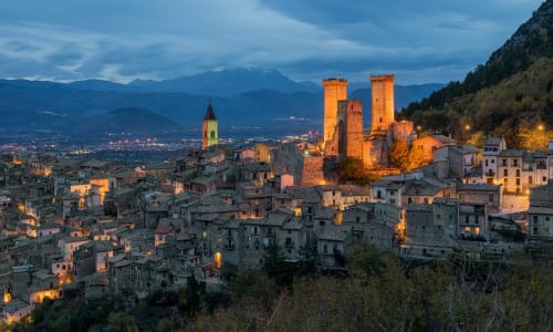 Borgo medievale di Sulmona al tramonto con torri illuminate, architettura storica e le montagne dell'Appennino sullo sfondo