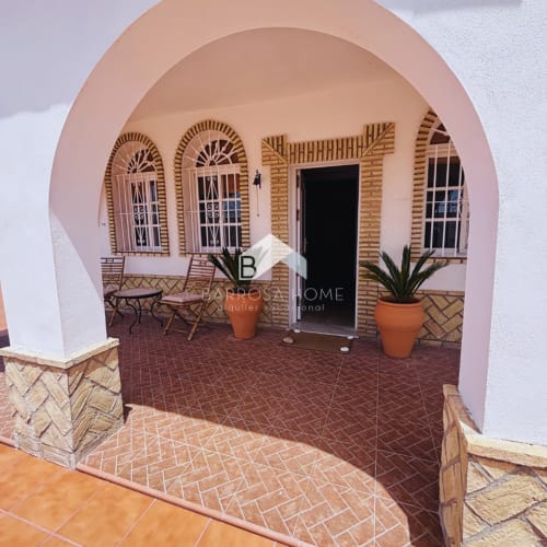 Entrada arqueada rosa a Villa Faro en Barrosa Home con patio de terracota, plantas en macetas y sillas de madera