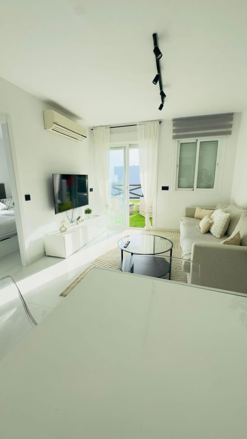 Sala de estar moderna en apartamento frente al mar con vistas al océano, TV de pantalla plana, sofá beige, mesa de cristal y acceso a terraza
