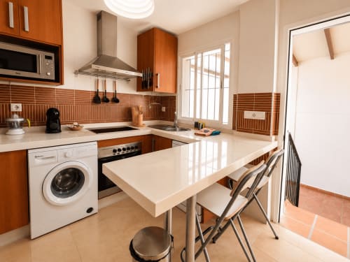 Cocina moderna con muebles de madera, encimeras blancas, mesa comedor y lavadora en apartamento Aroma de Poniente,