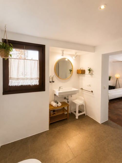 Bany modern de l'Hotel Mas Ribas amb mirall rodó, lavabo, estanteria de fusta i banqueta blanca accessible a Palamós