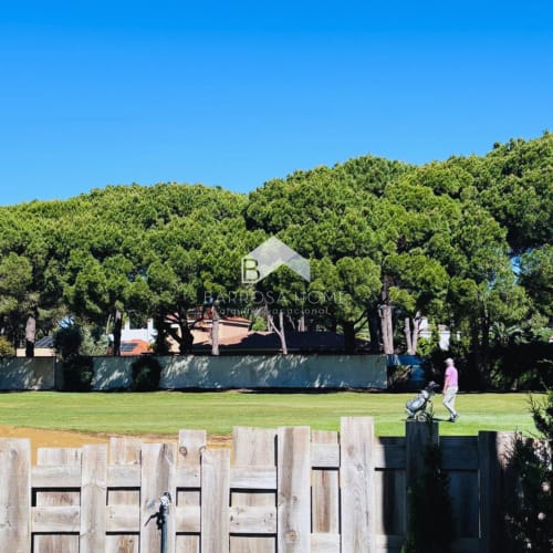 Villa Barrosa en Chiclana con frondoso jardín, césped cuidado, valla de madera y paisaje arbolado bajo cielo azul